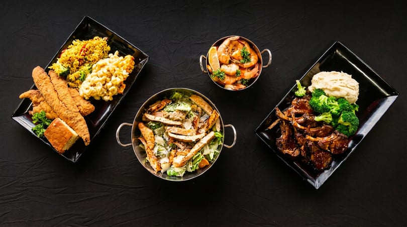 Top-down view of various dishes including fried fish, lamb chops, shrimp, and salads on a black background.