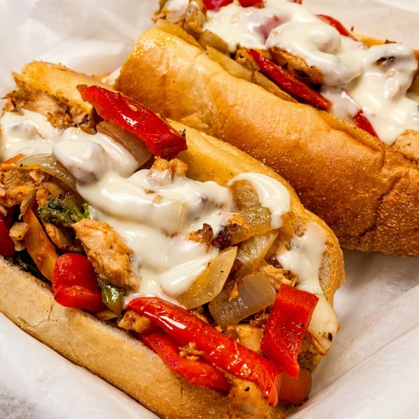 Two chicken cheesesteak sandwiches with melted cheese, red peppers, and onions in a white paper-lined tray.