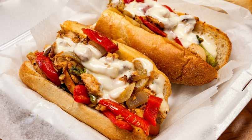 Two chicken cheesesteak sandwiches with melted cheese, red peppers, and onions in a white paper-lined tray.