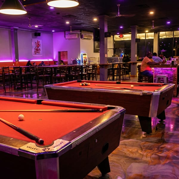 Two red-felt pool tables in a vibrant bar with purple lighting, patrons, and a long bar.