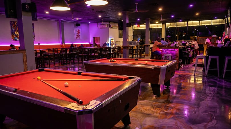 Two red-felt pool tables in a vibrant bar with purple lighting, patrons, and a long bar.