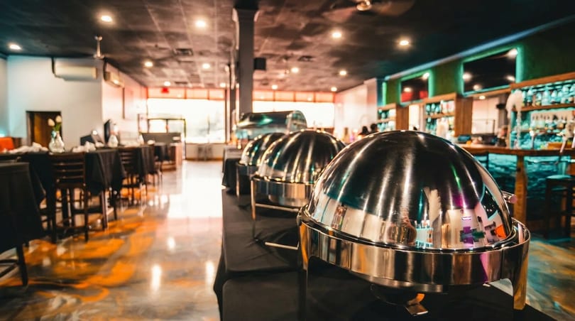 Buffet chafing dishes on a black-draped table in a restaurant with a bar and dining area.